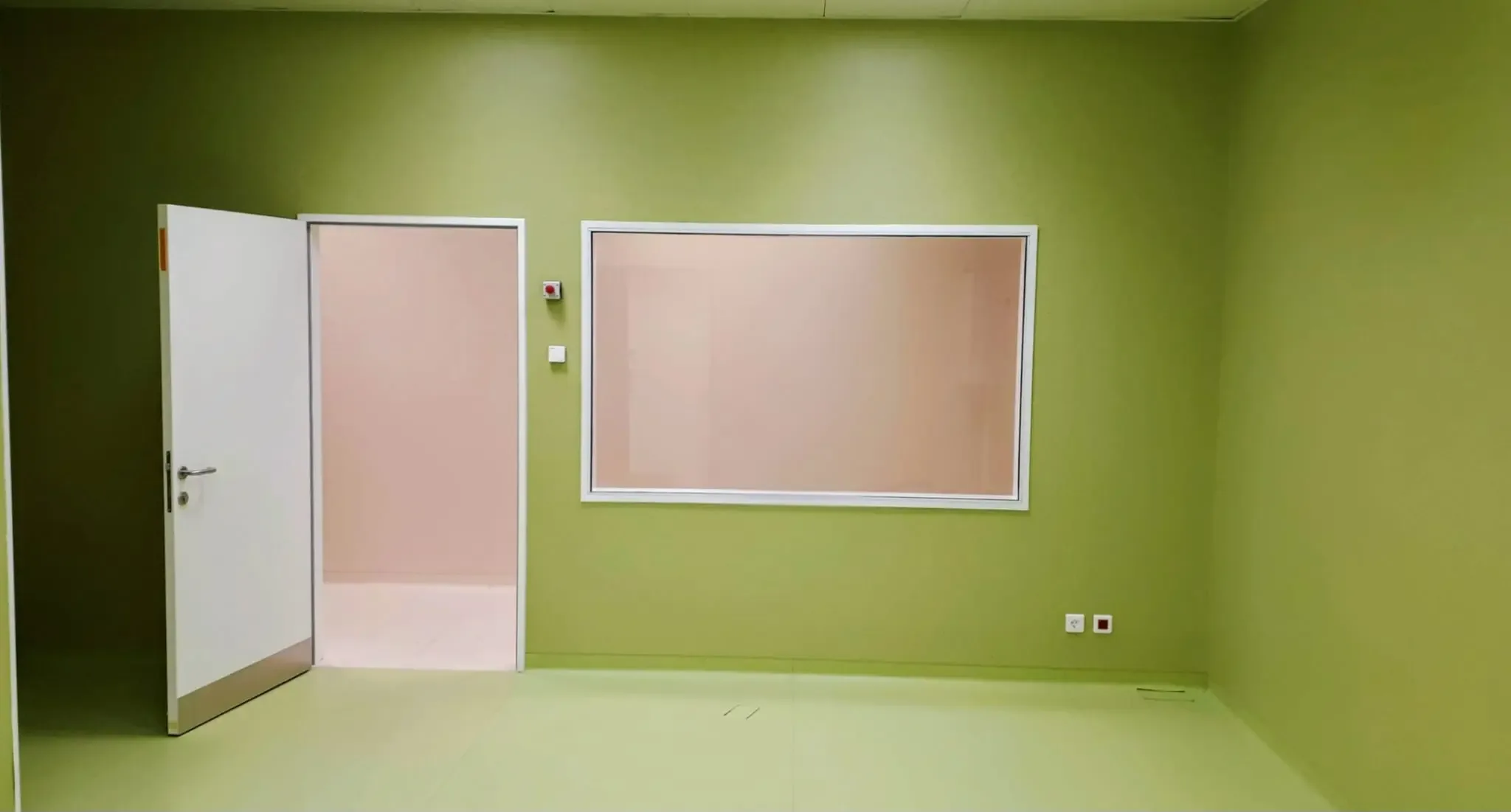 A room with green walls, an open door and protective lead glass in a white frame