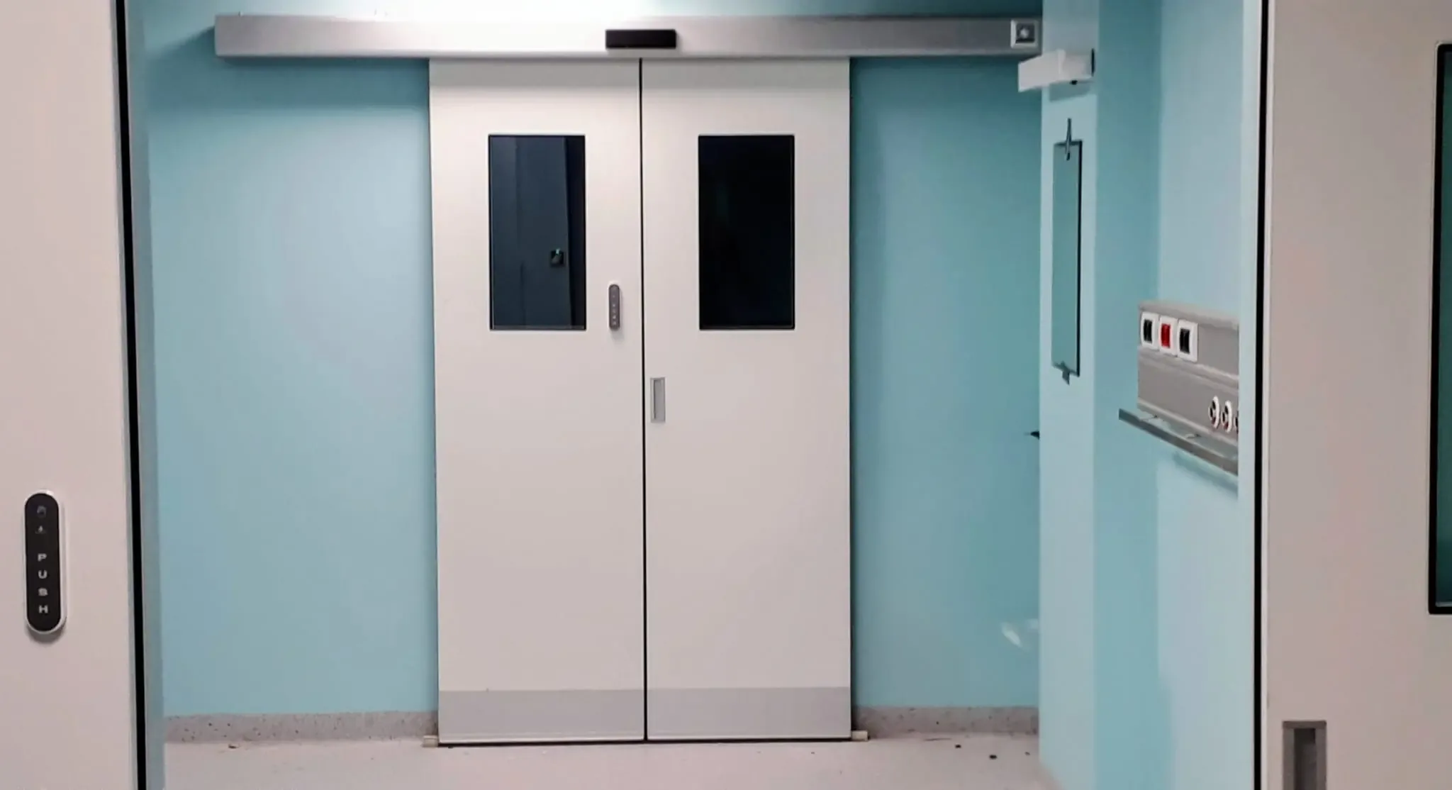 Double-leaf automatic sliding doors with narrow vision panels and a push-button opener, in a hospital corridor