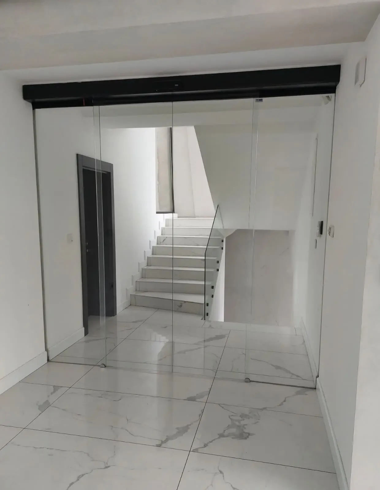 Full-glass automatic sliding doors with a drive in a black housing, in a lobby with a marble floor and staircase