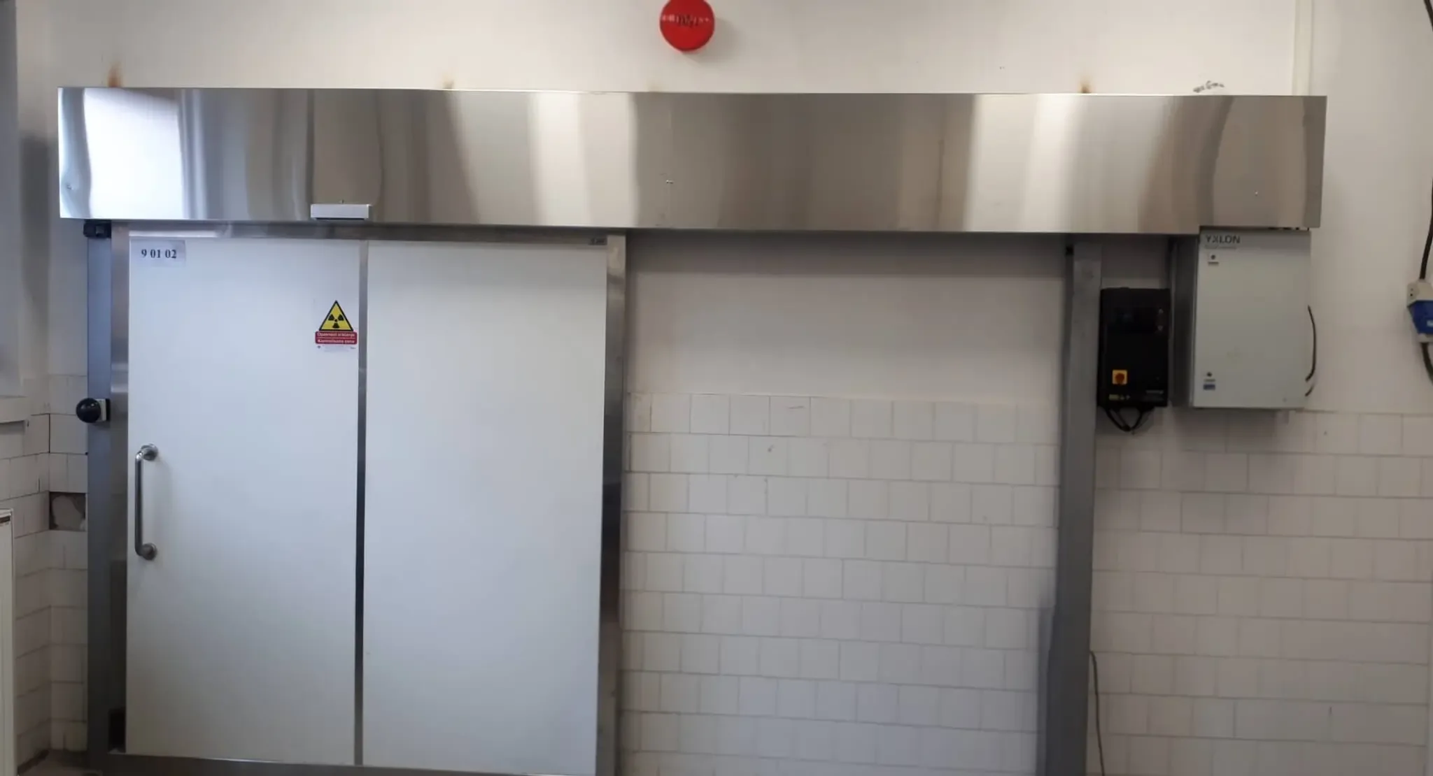 A massive sliding shielding door with a stainless steel drive housing, a red indicator light and a control cabinet