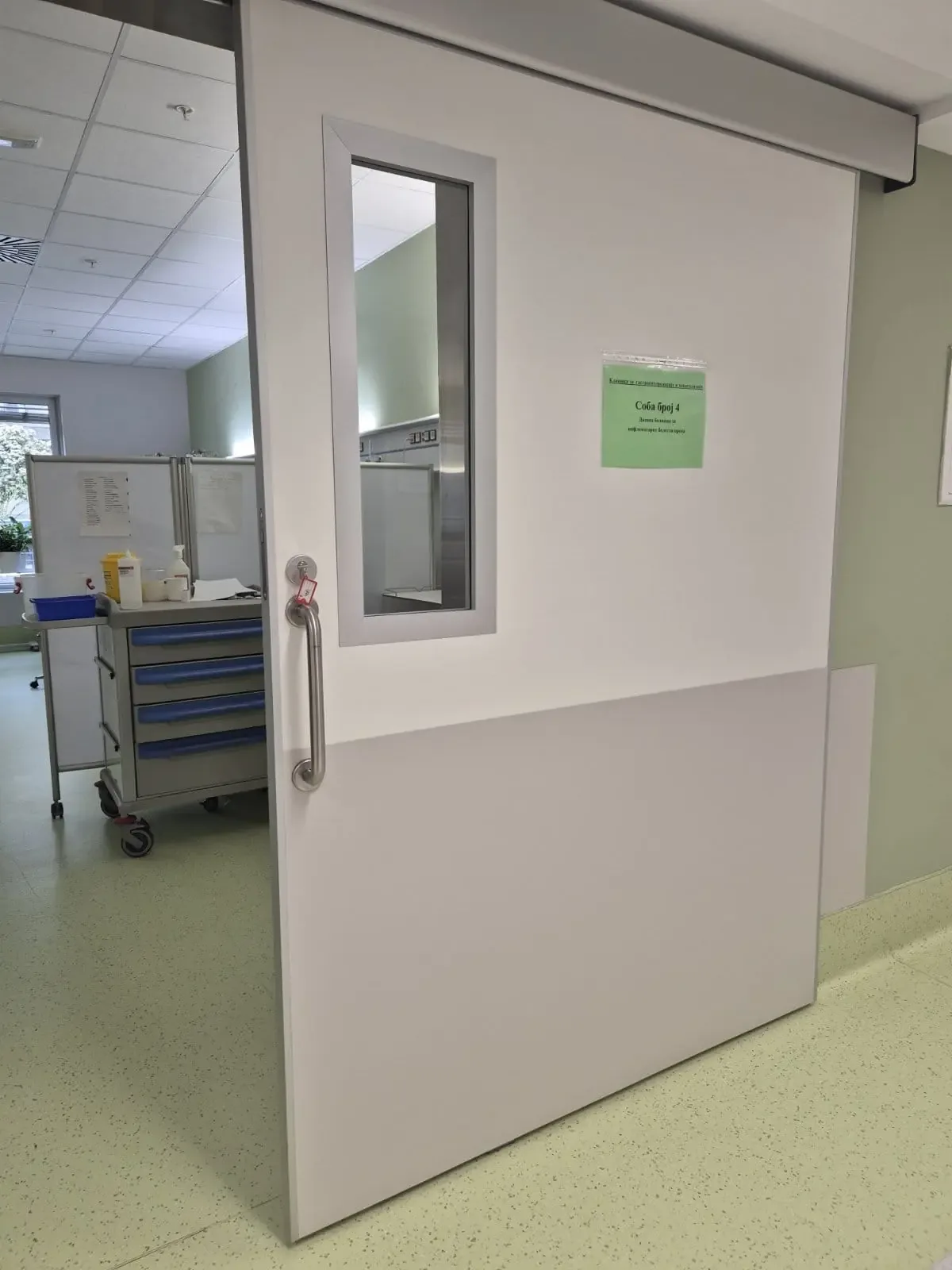 A ward door with a vision panel and a stainless steel handle, open onto a room with a medical trolley