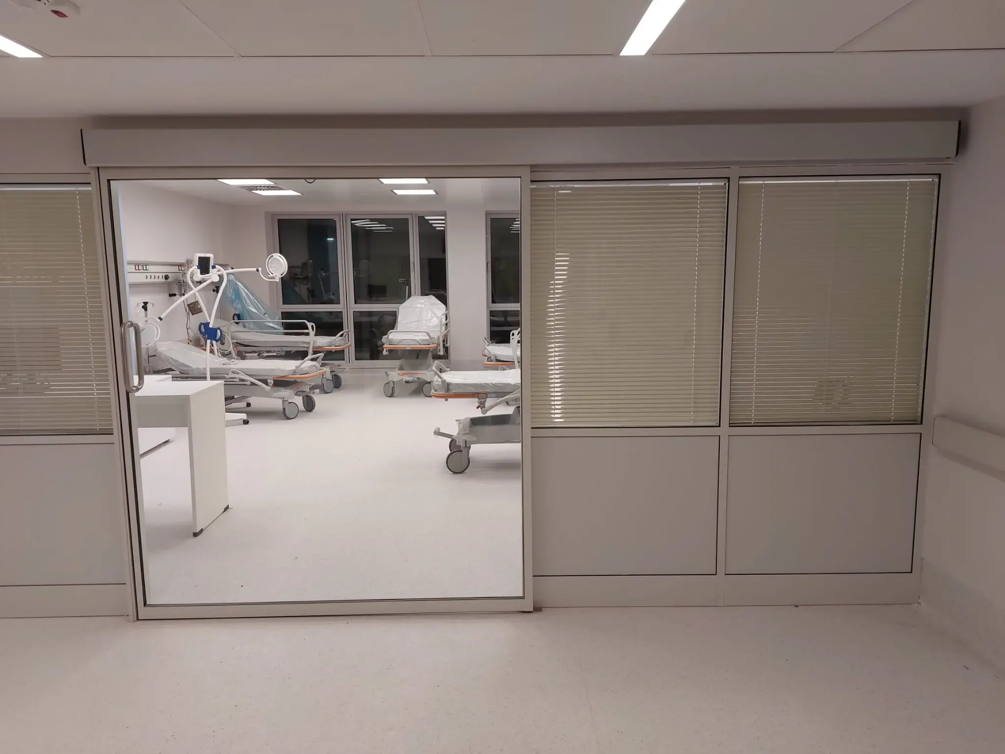 A glazed sliding partition with blinds onto an intensive care room with hospital beds