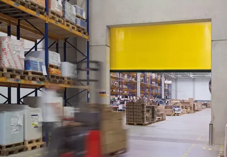 A high-speed door with a yellow curtain and a transparent panel in a warehouse with racking and a forklift
