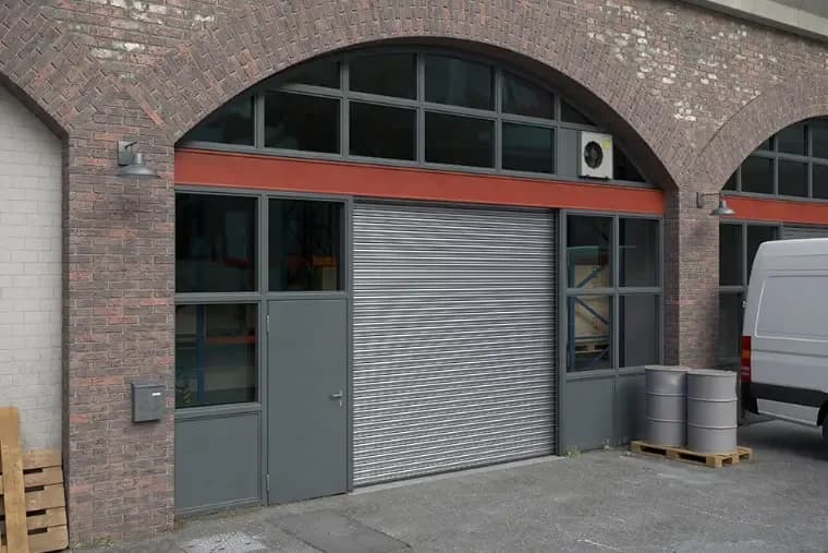 A roller shutter door and a side door in the arched opening of a brick building, with a van on the approach