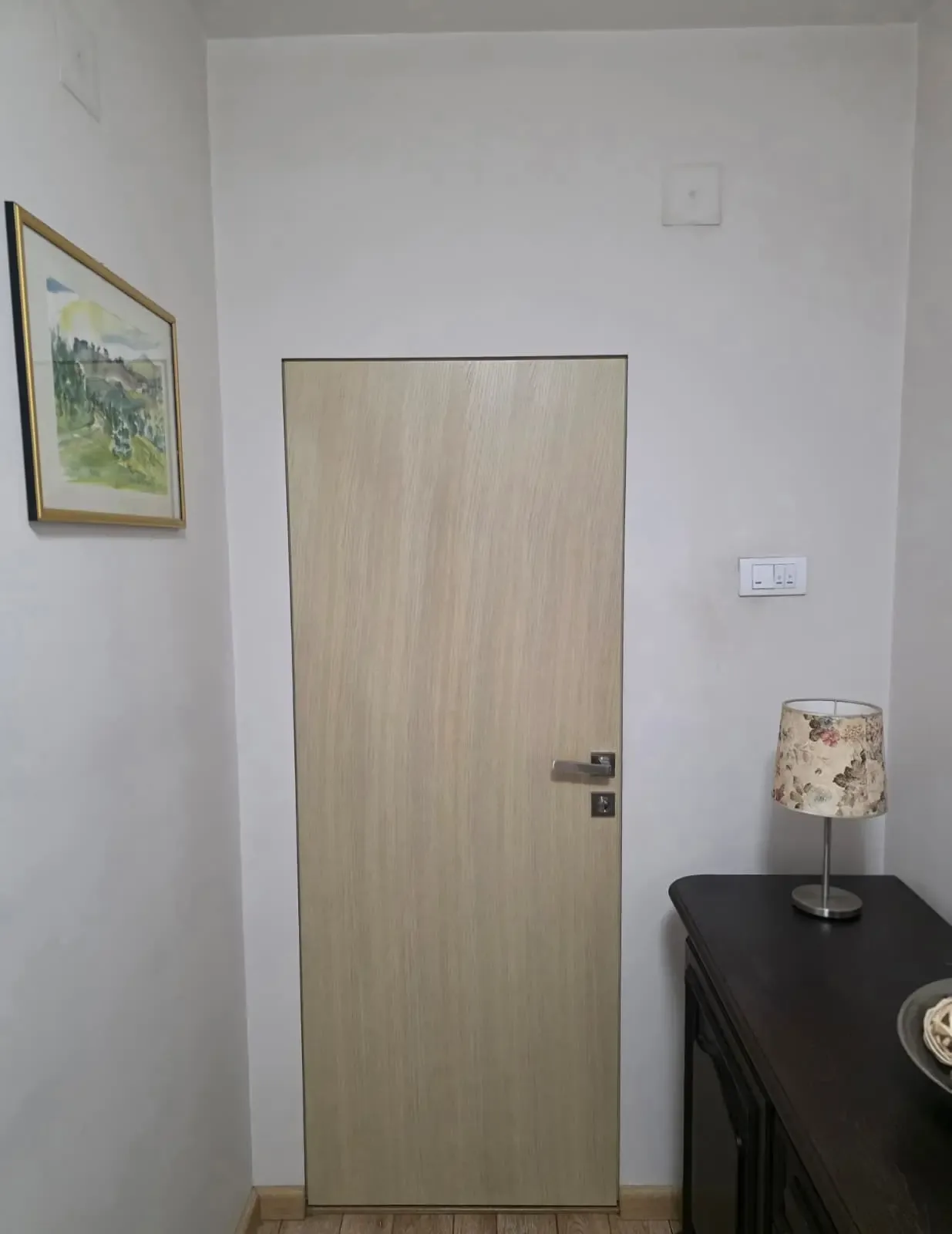 An interior door in a light timber finish in a residential hallway, beside a framed picture and a side table with a lamp
