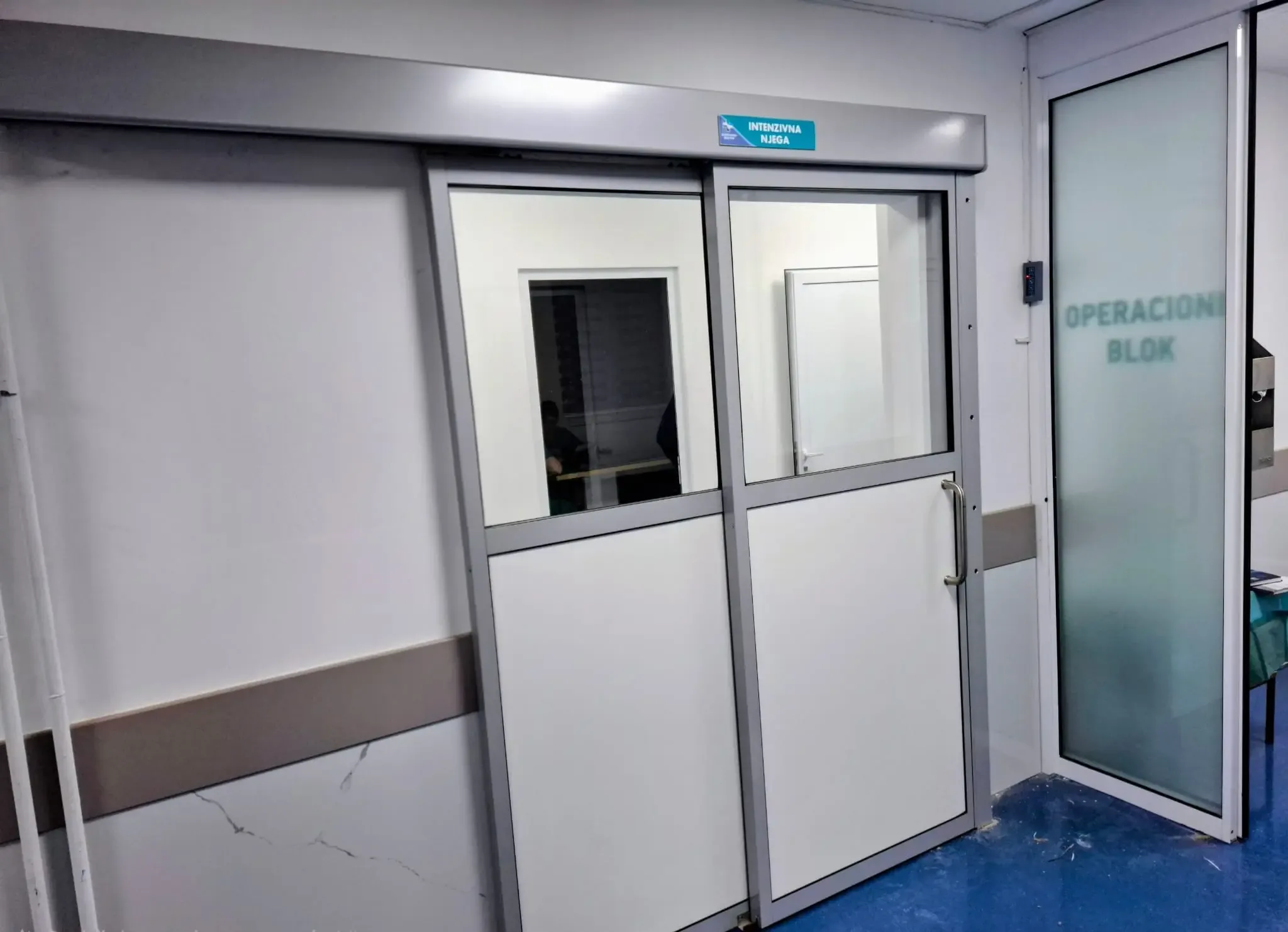 White double-leaf doors with vision panels and a glazed partition outside an operating suite